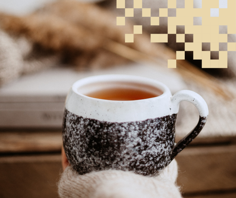 Käsi pitelemässä teekuppia, taustalla rekvisiittaa ja vaaleanruskeat pikselit.