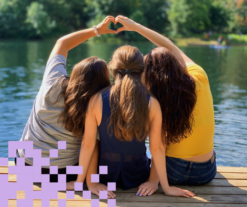 Kolme tyttöä laiturilla istumassa, kaksi muodostaa sydämen sormilla. Violetit pikselit alareunassa.
