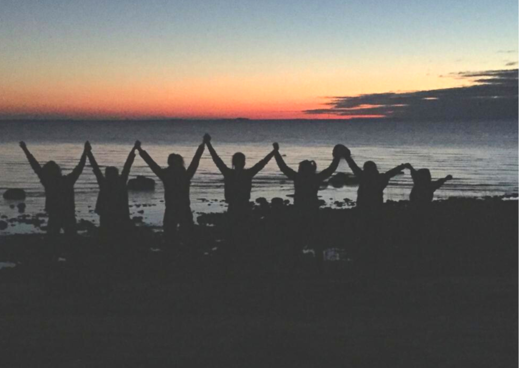 Kuvassa useat henkilöt seisovat lähellä toisiaan auringonlaskun edessä meren rannalla pitäen toisiaan kädestä kiinni kohotetuin käsin. 