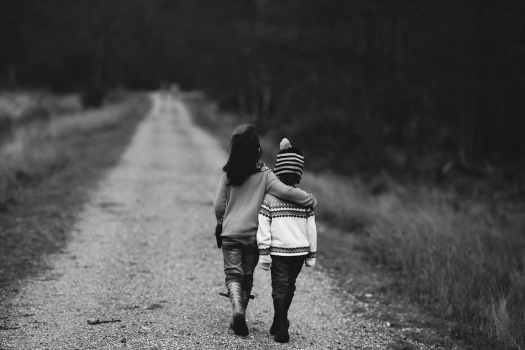 Mustavalkoisessa kuvassa kaksi lasta kävelee tietä pitkin. Toinen pitää toista kainalossa