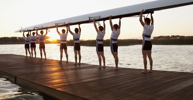 Urheilujoukkue kannattelee suurta kanoottia päidensä päällä, seisovat laiturilla auringon noustessa.